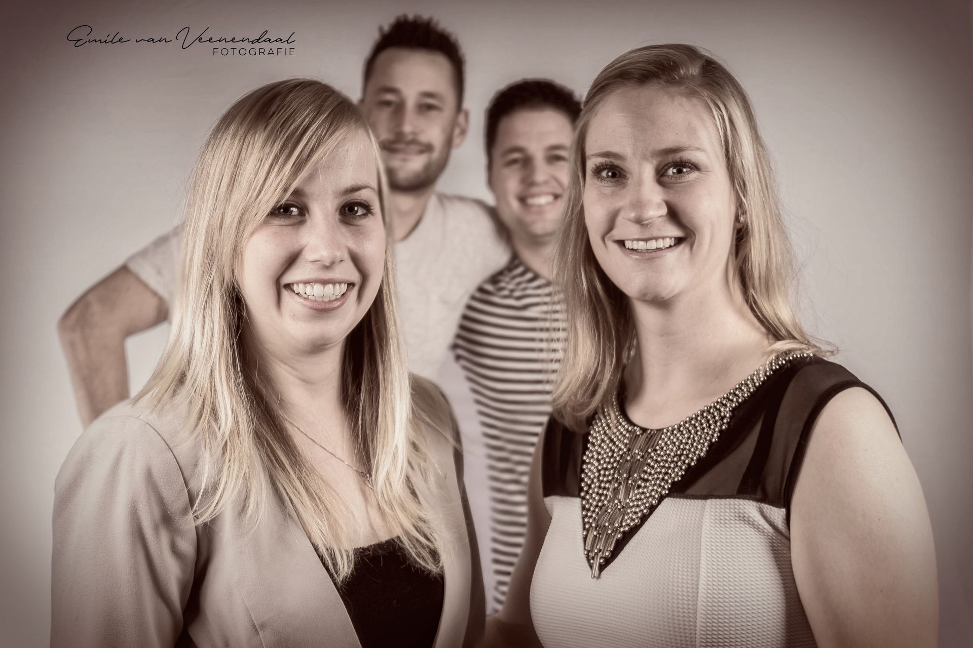 Vier lachende mensen poseren binnen, met twee vrouwen vooraan en twee mannen op de achtergrond.