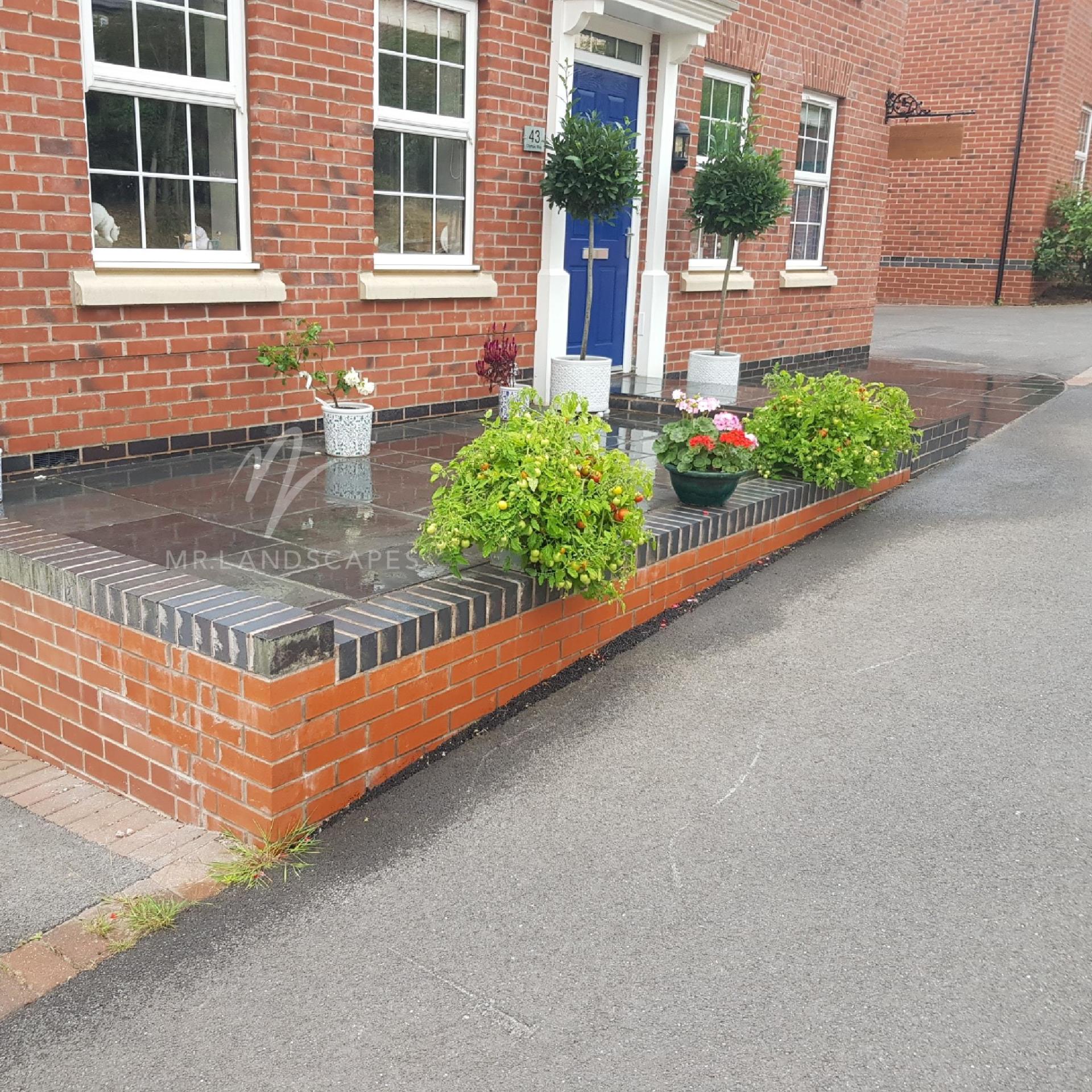 Brick townhouse entrance with potted plants and a small raised flower bed by the sidewalk