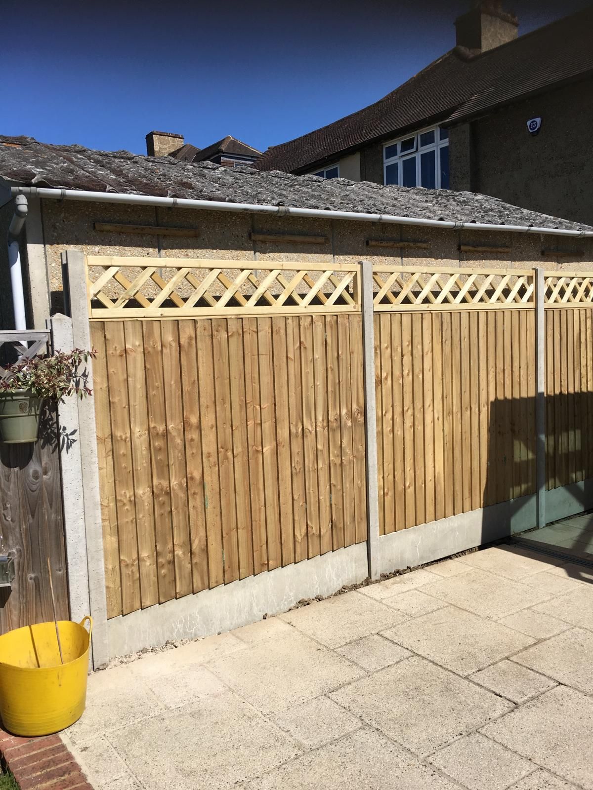 Wooden backyard fence with lattice top beside a paved patio and yellow bucket
