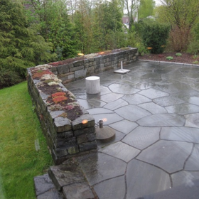 Stone patio with low retaining wall, wet flagstones, and small white stools beside a grassy yard.