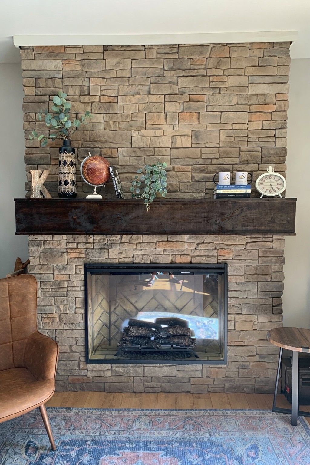 A stone fireplace with a dark wooden mantel, topped with a globe, vase, and decor, beside a leather armchair.