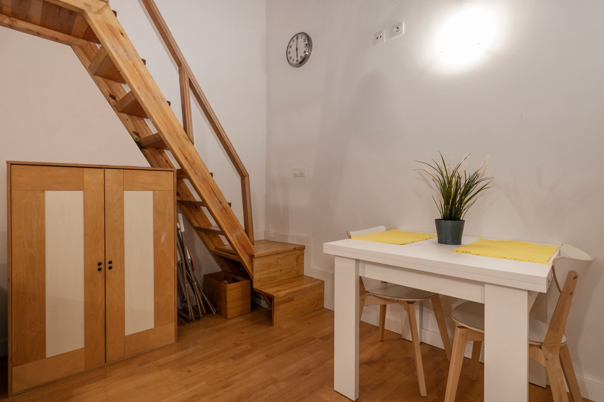 Small dining nook with wooden stairs, a white table with plant, and a cabinet under a wall clock.