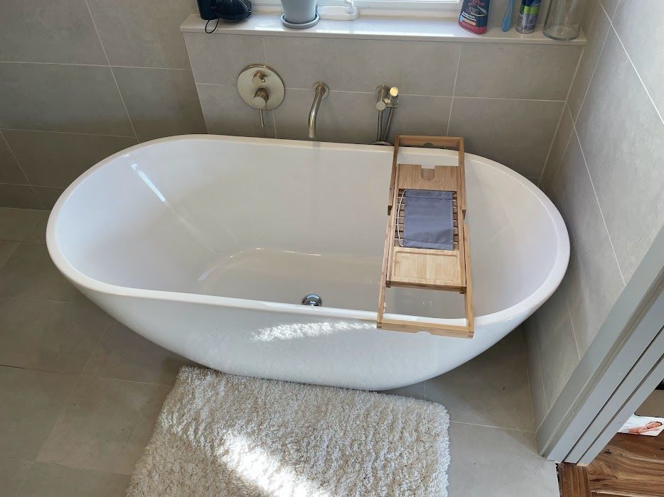 White bathtub in a tiled bathroom with a small wooden tray across the rim and a bath mat nearby