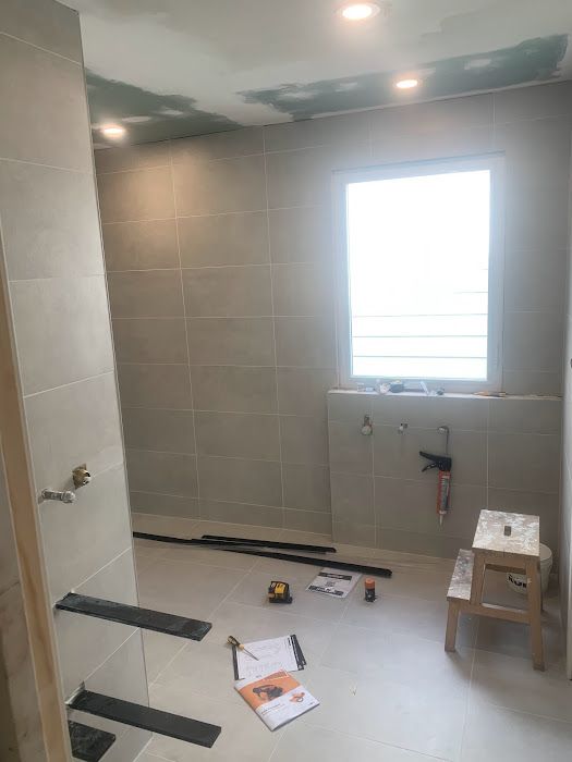 Bathroom under construction; light gray tiled walls, small window, tools on floor, unfinished ceiling.