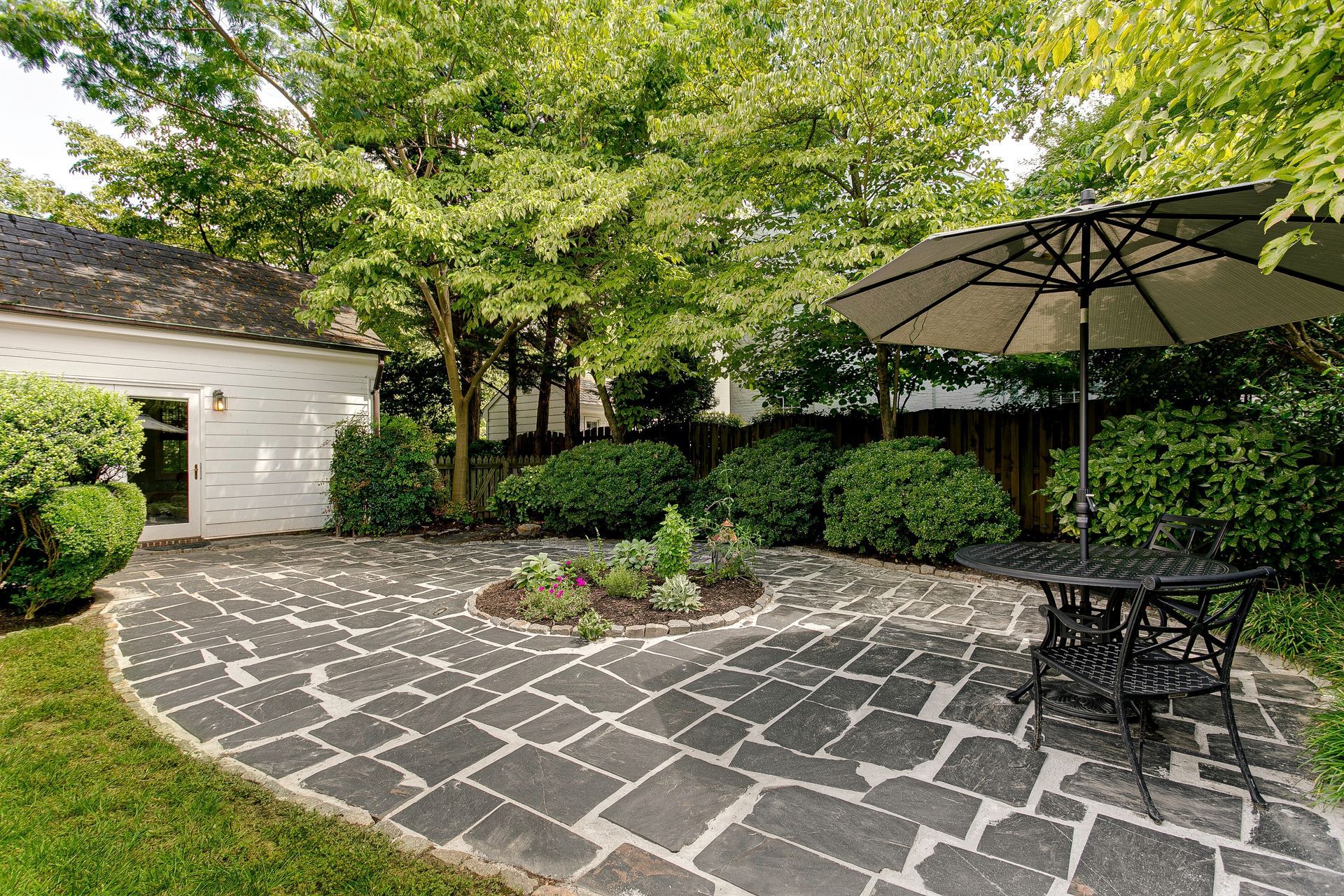 Stone patio with black table and umbrella beside a garage and lush green landscaping