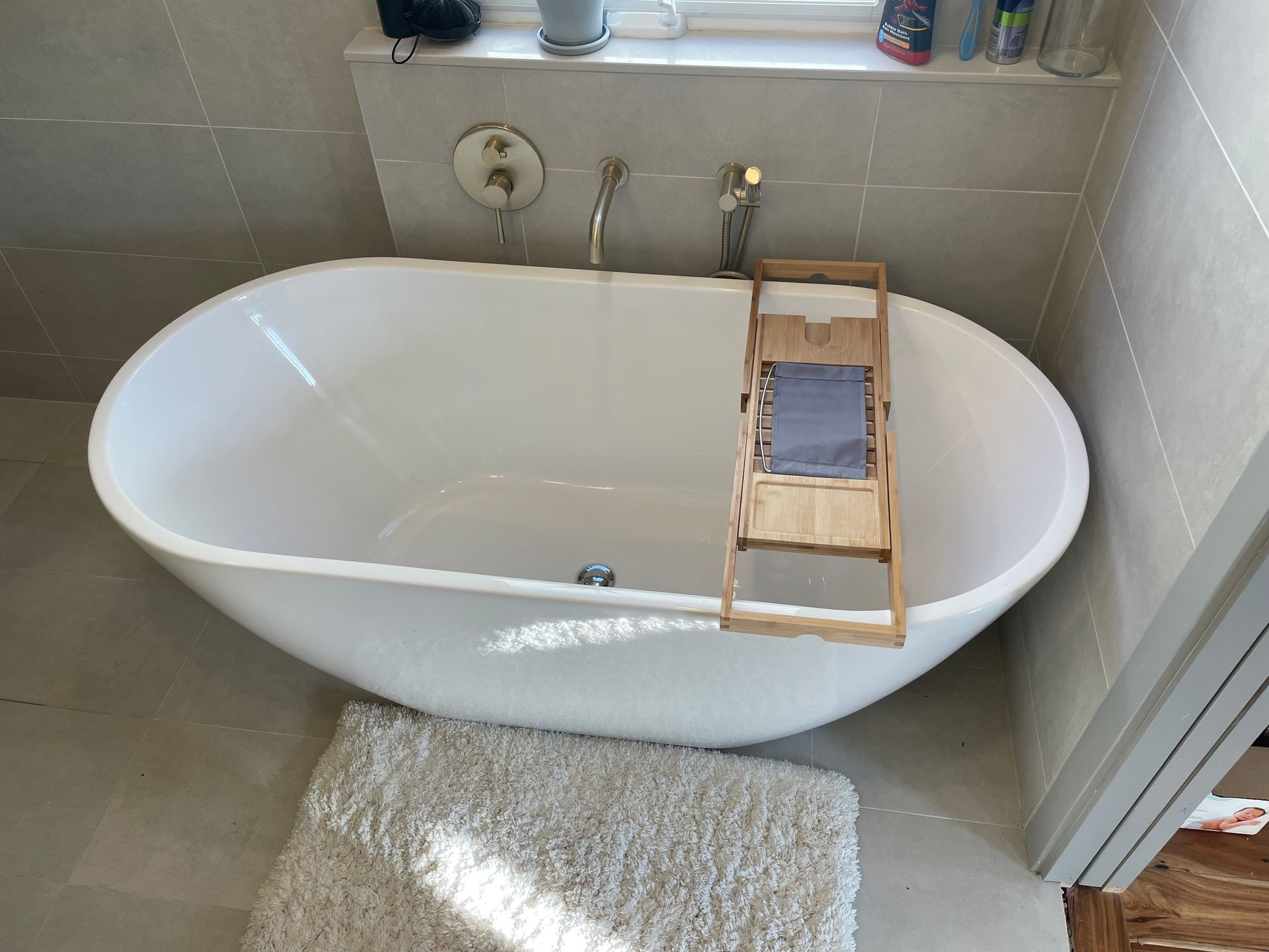 Oval white bathtub in a bathroom, with a bamboo tray holding a towel. A white rug is in front.