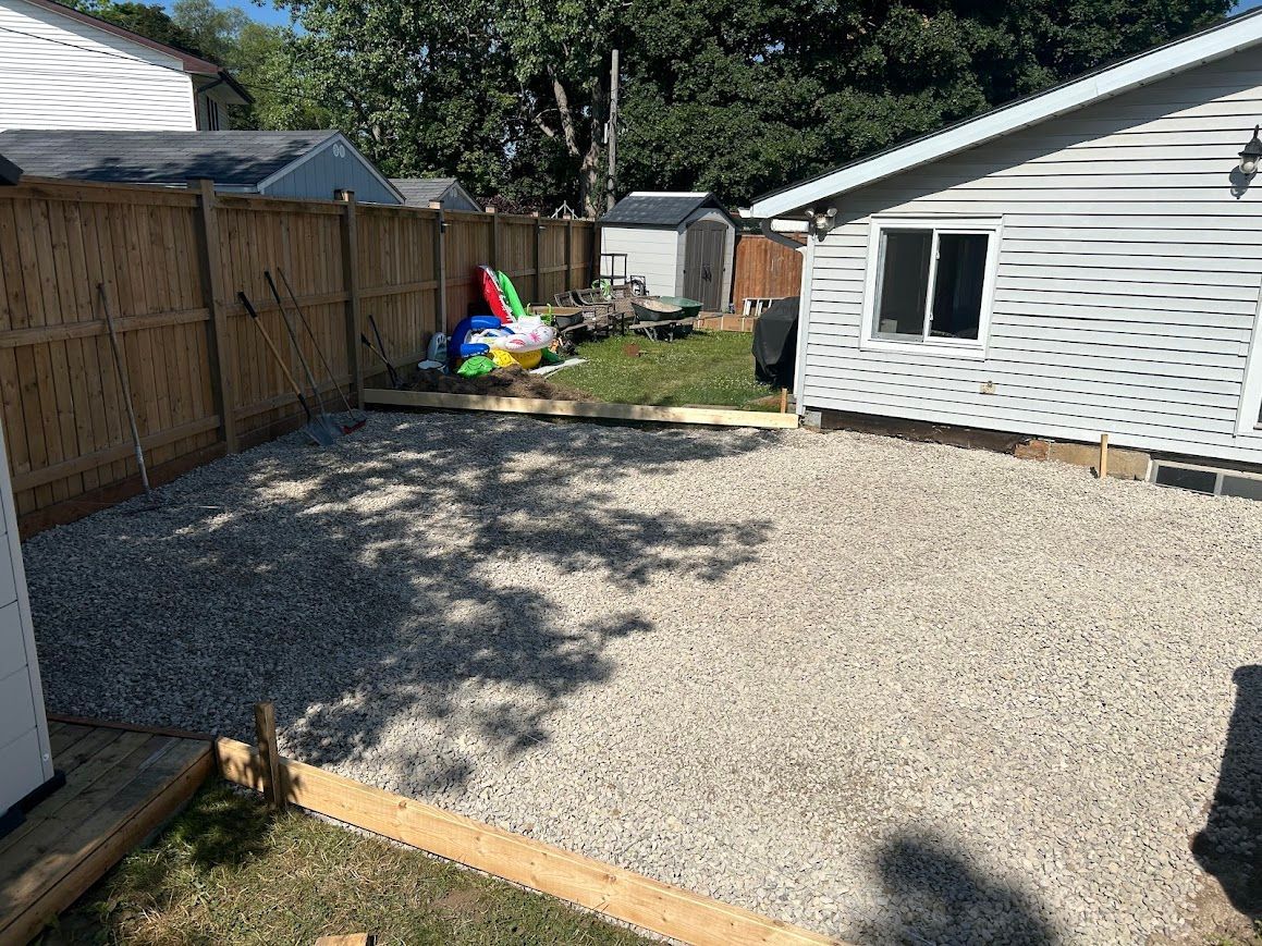Gravel-covered backyard with wooden fence and house. A few toys are near the grassy area.