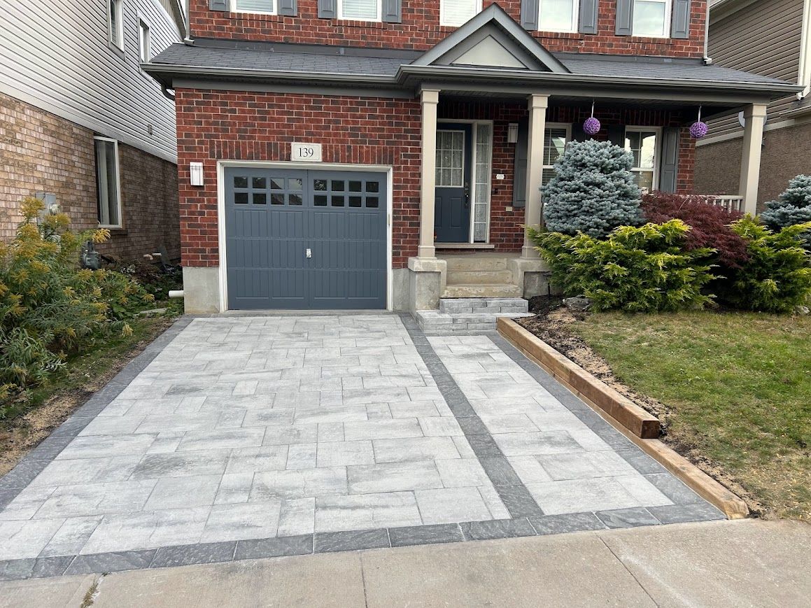 Brick house with gray paved driveway and walkway leading to the front door, with a garage door and landscaping.