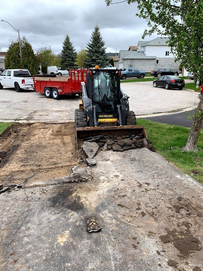 Skid steer loader removing pavement on a residential driveway. Red trailer and parked vehicles in background.