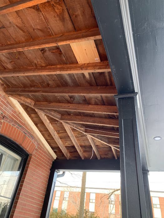 View of a porch ceiling with wooden planks and supports, next to a black column and brick wall.