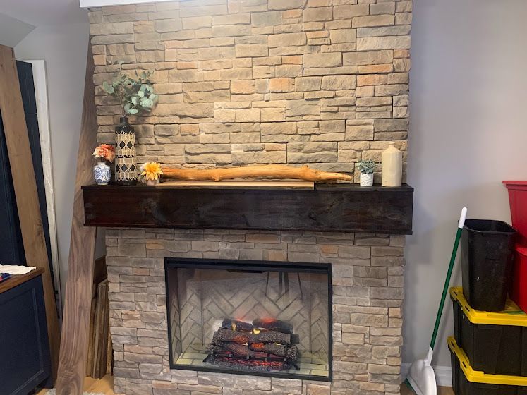 A stone fireplace with a dark wooden mantel, decorative items on top, and a hearth inside a room under construction.