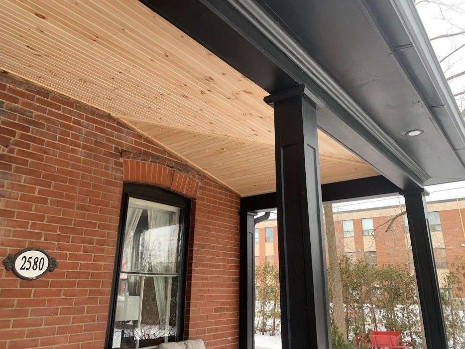 A covered porch with wood ceiling, supported by black pillars, next to a brick building.