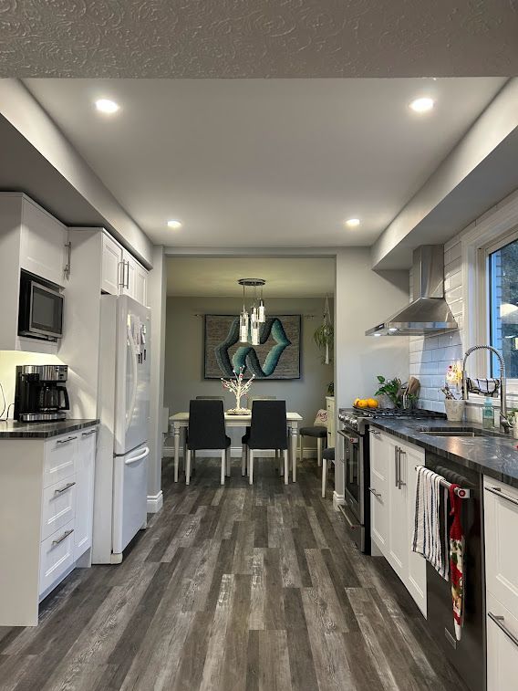 Modern kitchen with white cabinets, dark gray countertops, and wood-look flooring. Leads to a dining area.