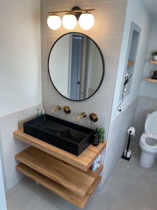 Modern bathroom with black sink, round mirror, wood shelves, gold fixtures, and gray tile.
