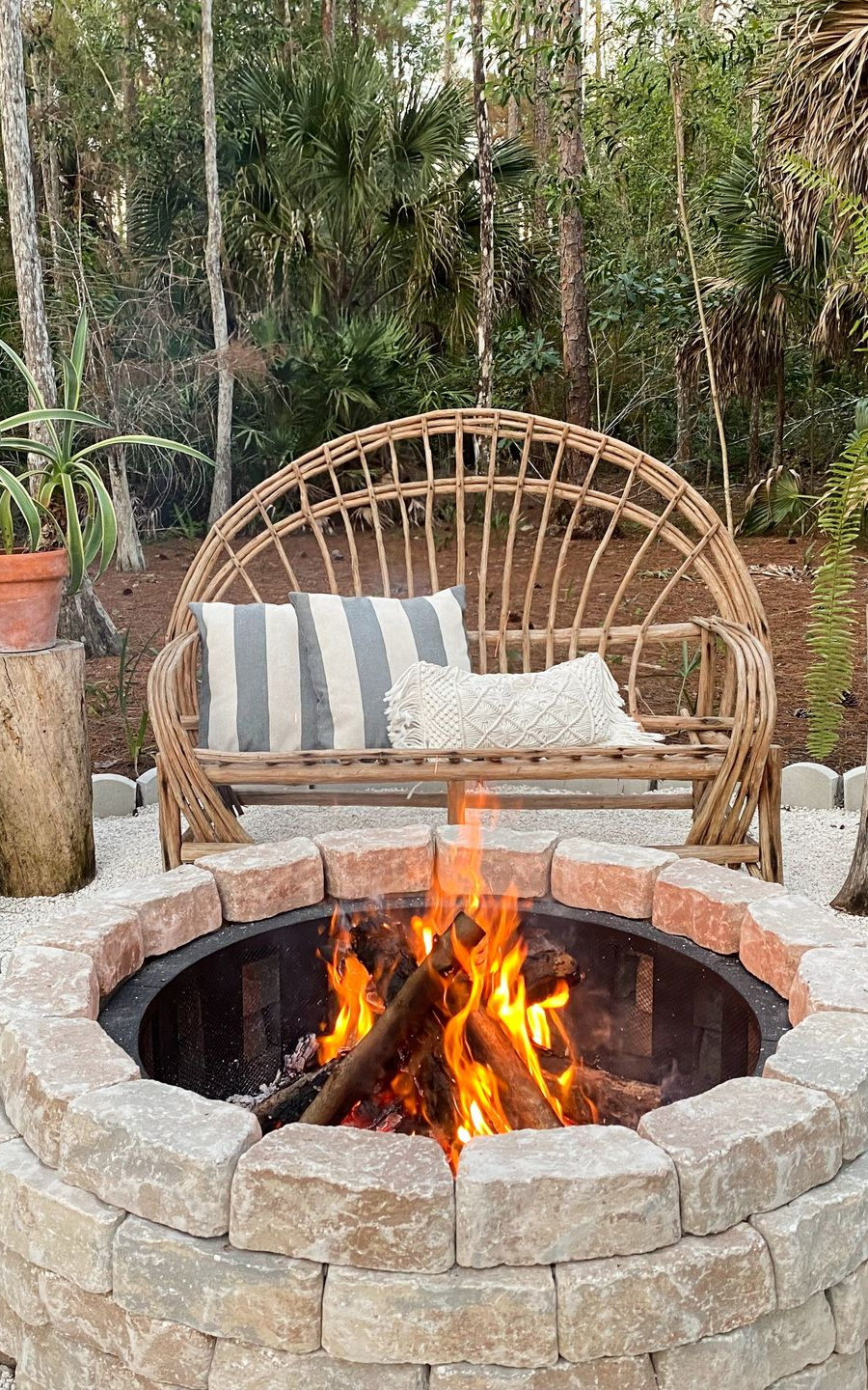 A fire pit with burning wood logs sits in front of a curved, light-brown wicker bench with striped and textured pillows.