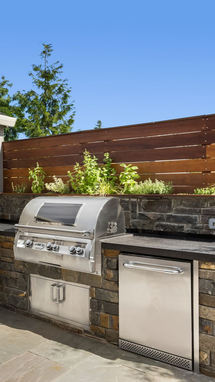 Outdoor kitchen featuring a stainless steel grill and mini-fridge set into a stone counter against a wood slat fence.