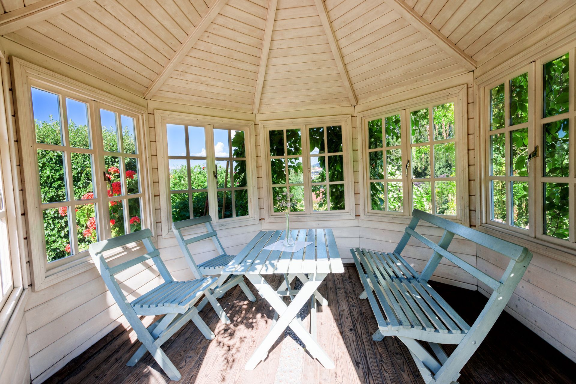 A light blue wooden table and chairs sit inside a bright, white-paneled gazebo with large windows looking out on greenery.