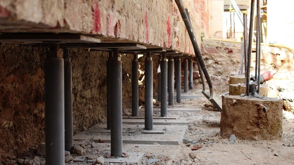 Underside of a damaged building supported by black steel posts, with rubble and a dirt trench beside it