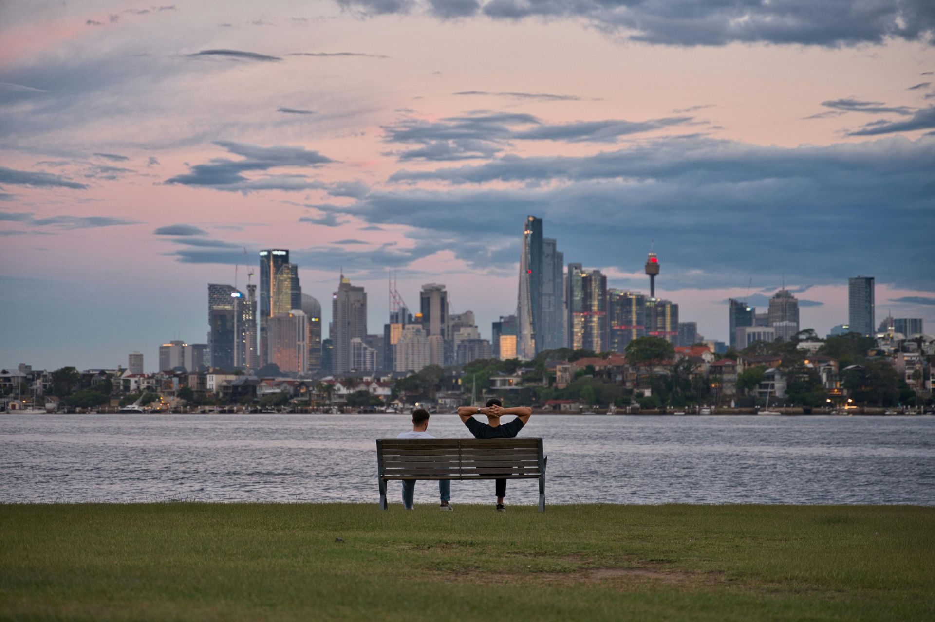 Person sitting on a bench by the water, with a city skyline at dusk in the background