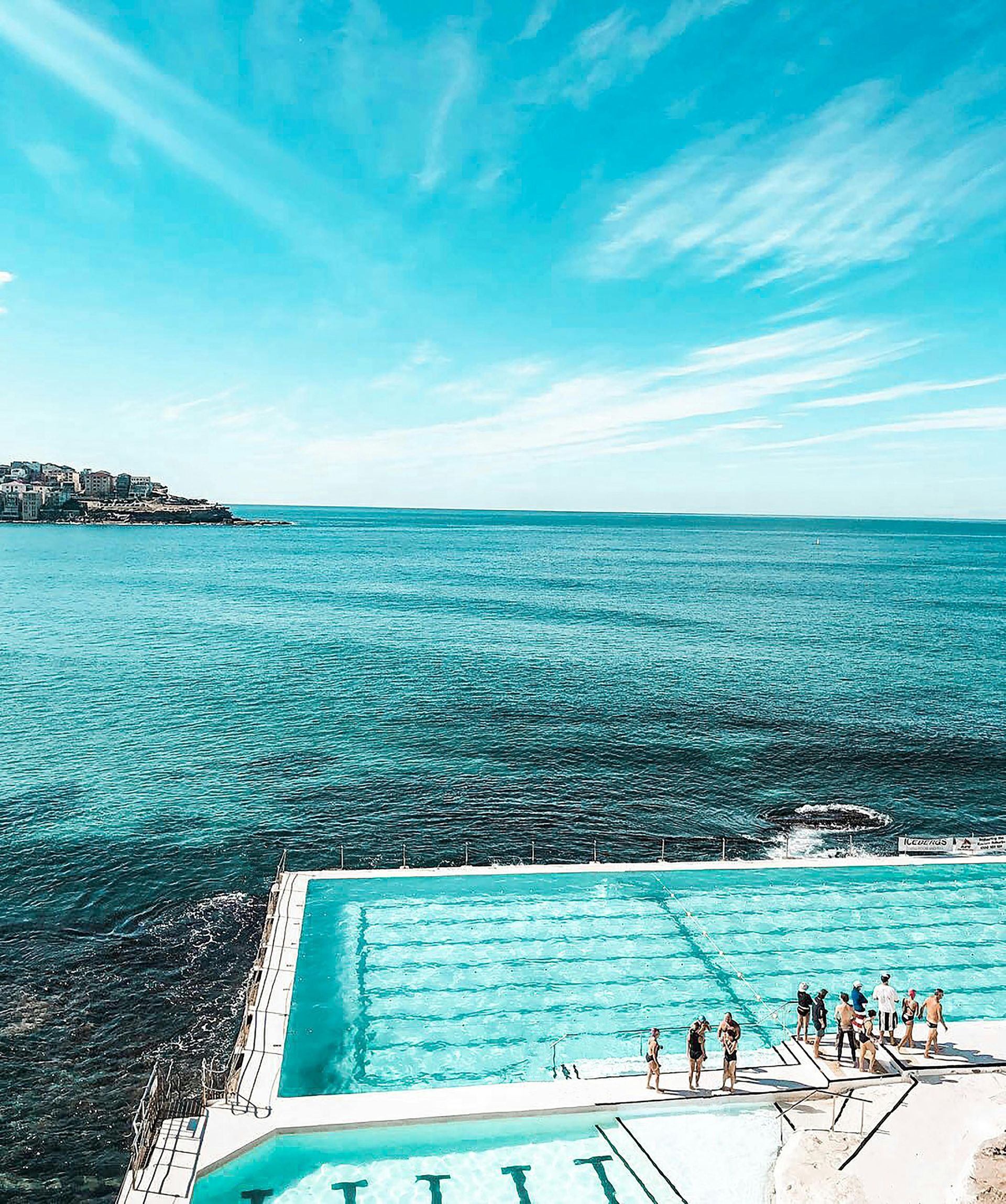 Turquoise seaside pool beside a rocky shore, with swimmers and a bright blue sky beyond the open water