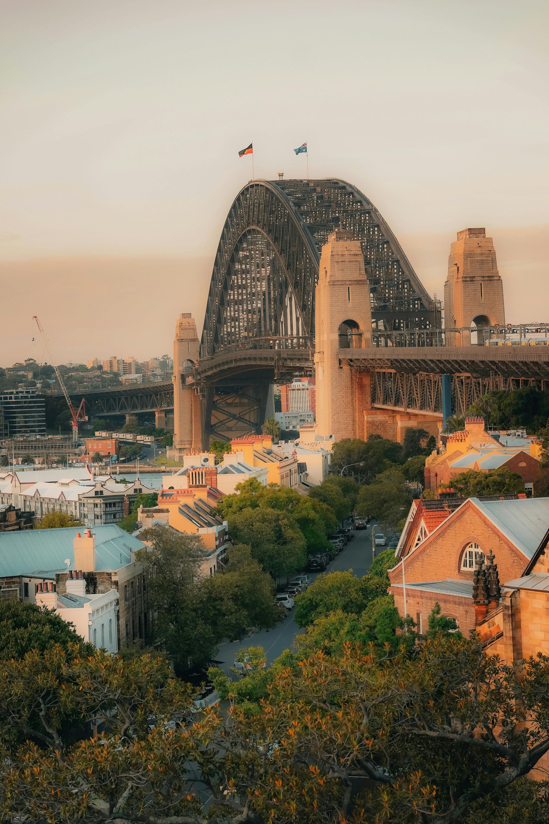 Sydney Harbour Bridge at sunset above city buildings and trees, with two flags on top