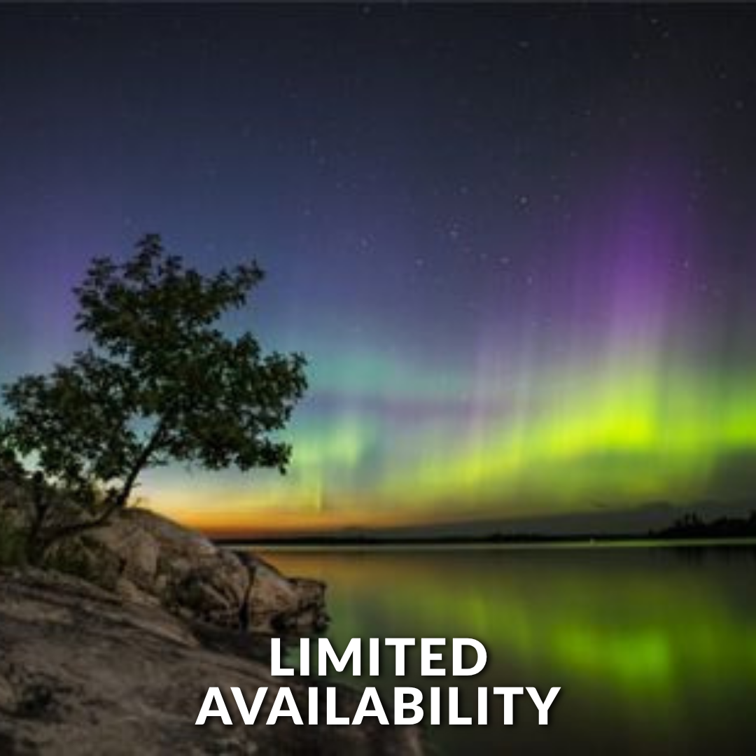 Vibrant green and purple aurora borealis illuminates the night sky above a calm, reflective lake and rocky shoreline.