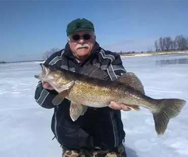 A person in a winter jacket and cap holds a large walleye fish while standing on a frozen, snow-covered lake.