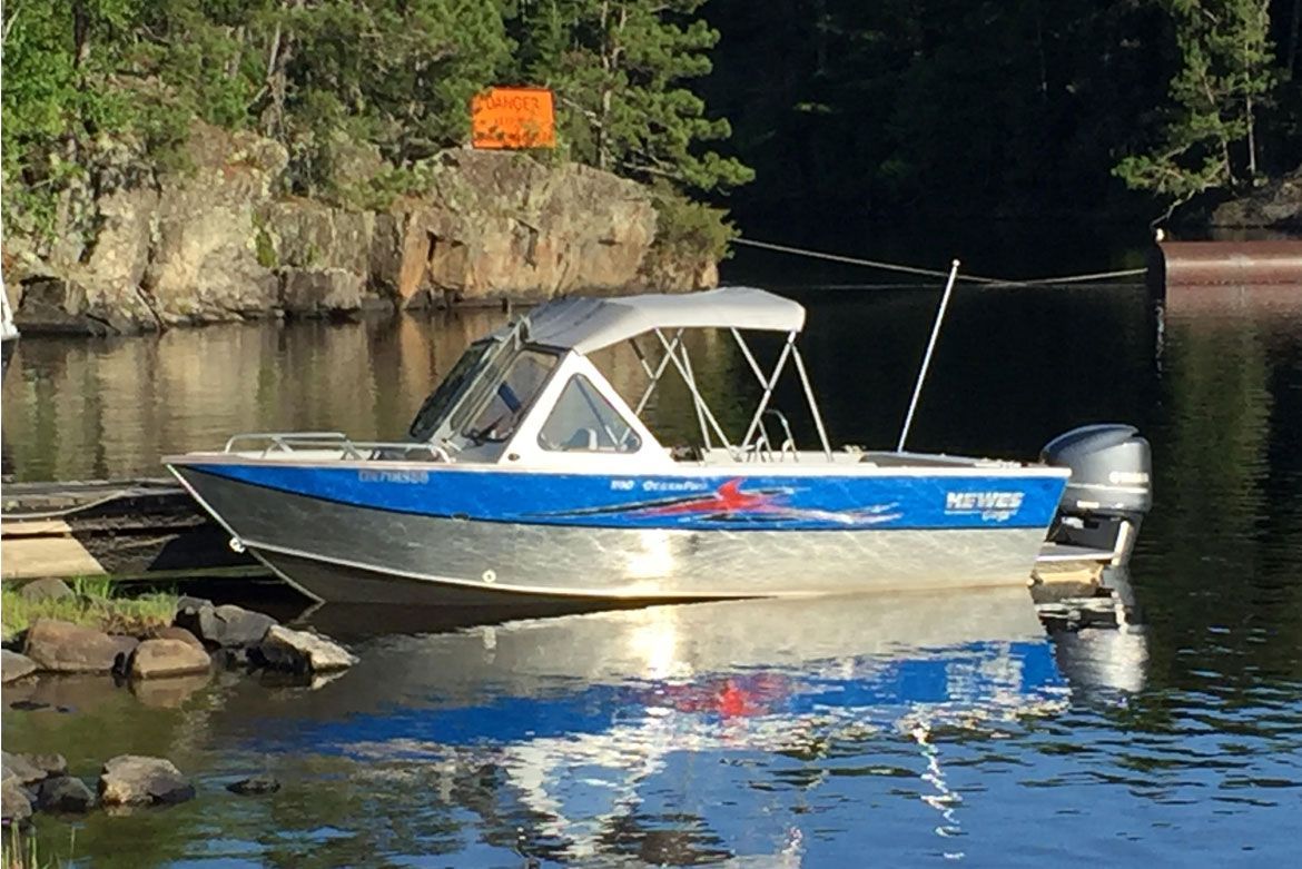 A blue and silver aluminum fishing boat with a canvas top docked at a rocky shoreline near a forest.