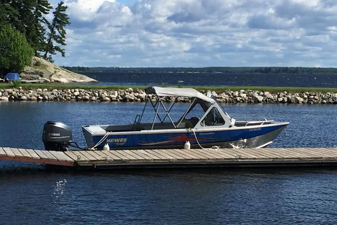 A blue and silver aluminum fishing boat with a canopy top docked at a wooden pier on a lake.