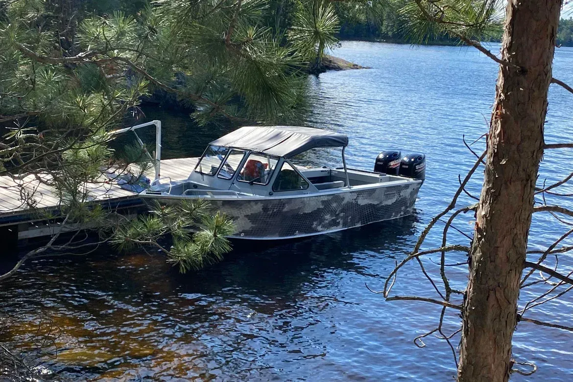 A camouflaged motorboat with twin engines is docked at a wooden pier on a sunny lake, framed by pine trees.