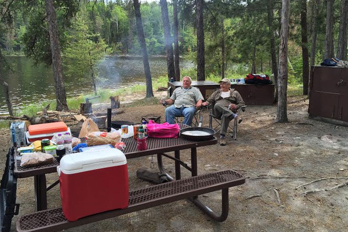 Two people sit in camp chairs by a picnic table laden with food and a red cooler, located in a wooded area near a lake.