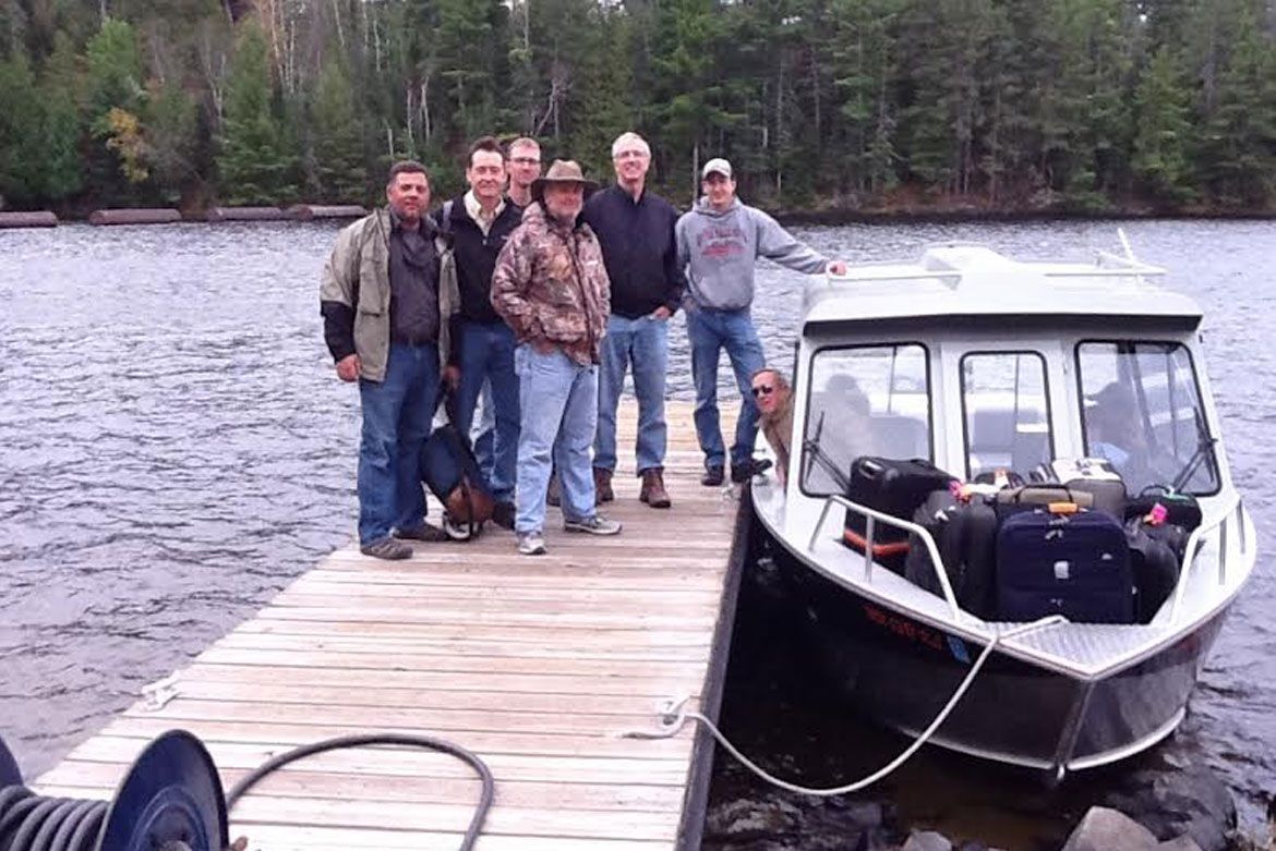 Six people stand on a wooden dock next to a boat loaded with bags, with a forest and lake in the background.