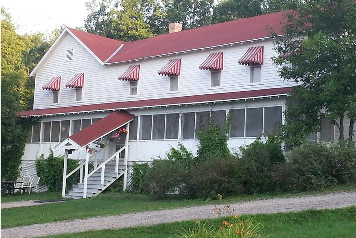 A white two-story house with a red roof, striped awnings, and a covered porch, set against a background of trees.