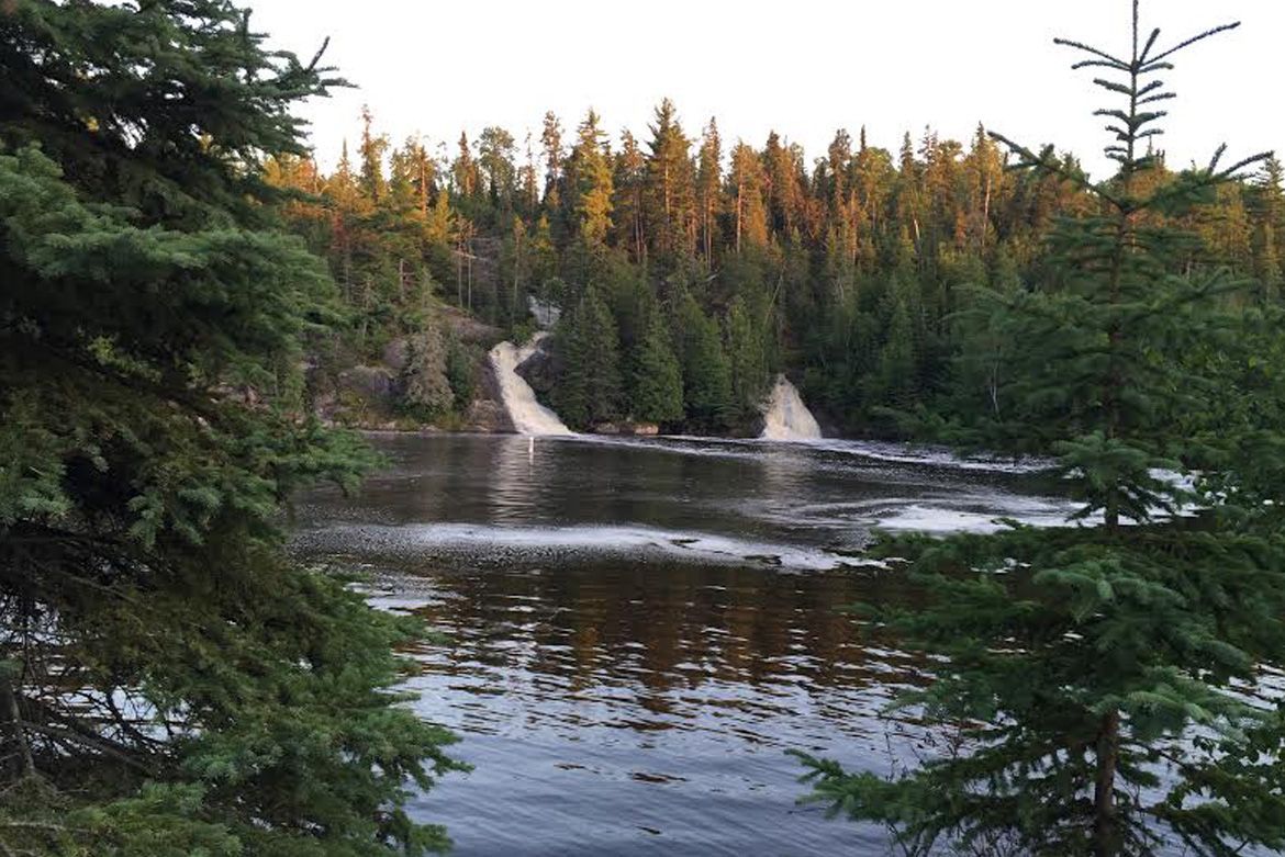 Two waterfalls cascade into a dark, calm lake surrounded by dense evergreen trees at sunset.
