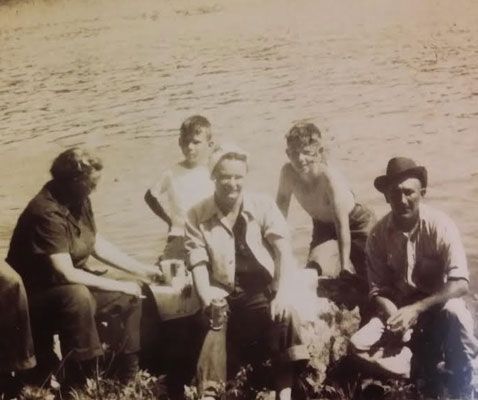 Group of people sitting and standing near a body of water, posed for a sepia-toned outdoor photograph.