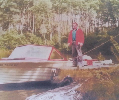 A person in a red jacket stands on a dock next to a boat near a forest.