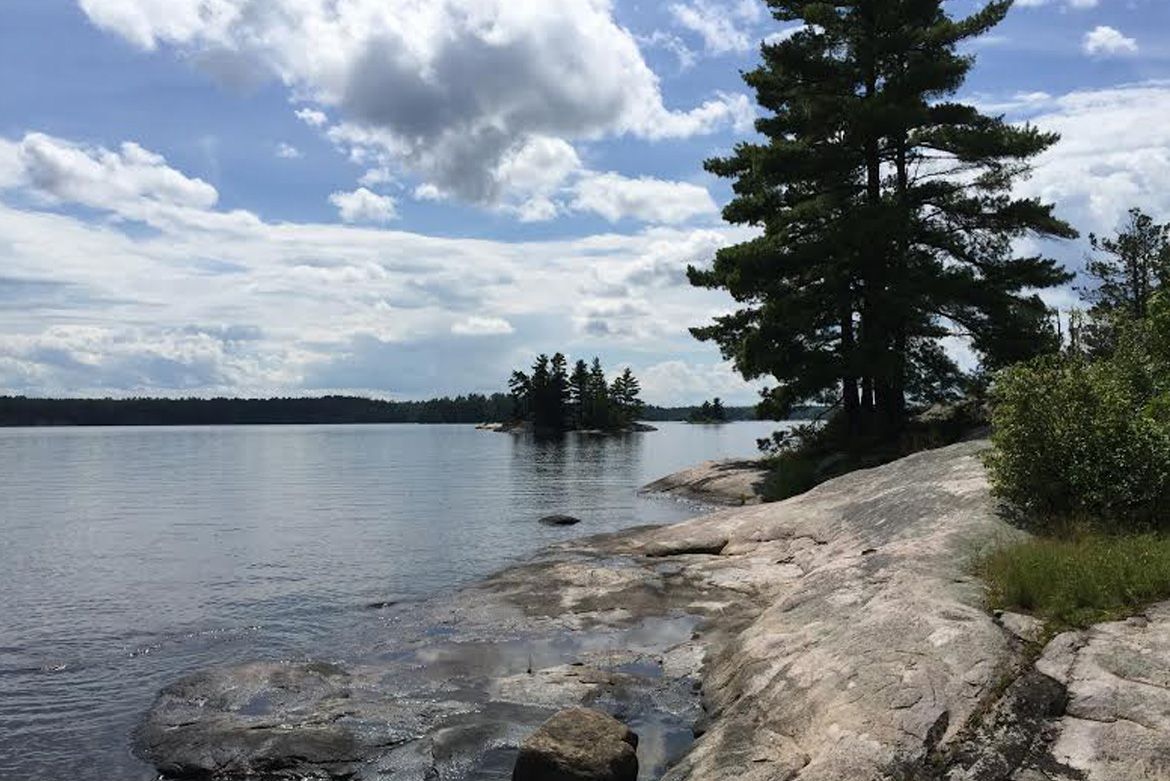 A calm lake under a cloudy sky with smooth rocky shores, pine trees, and a distant island.