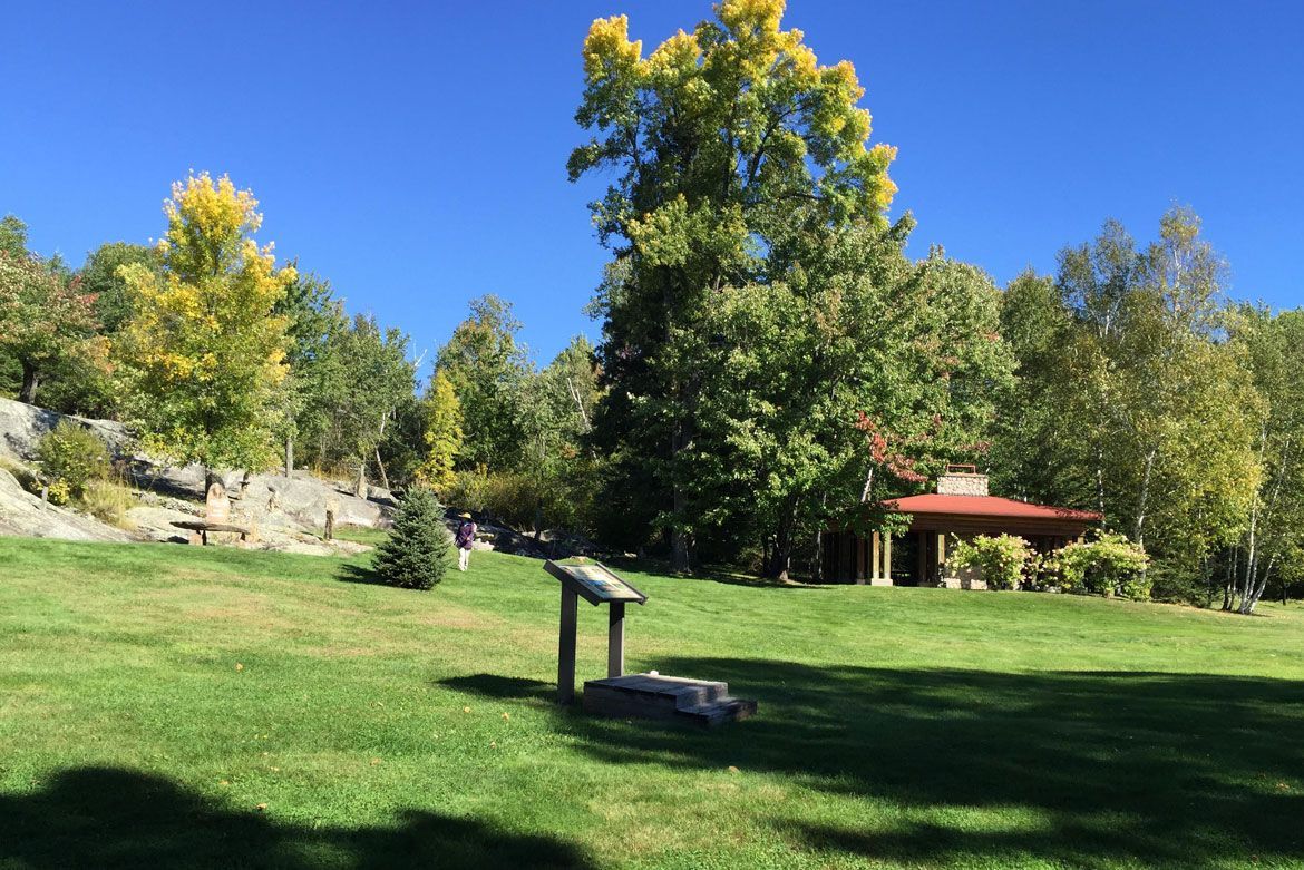 A grassy park with a trail sign in the foreground, trees with autumn foliage, and a small building under a clear blue sky.