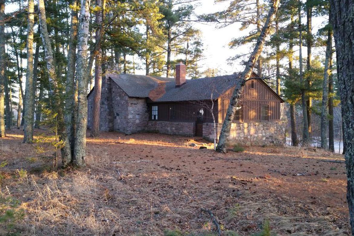 A stone and wood cabin nestled in a wooded clearing with tall pine trees during the late afternoon sun.