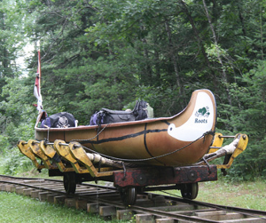A tan Roots-branded canoe rests on a small, yellow-framed wooden railway car in a wooded area.