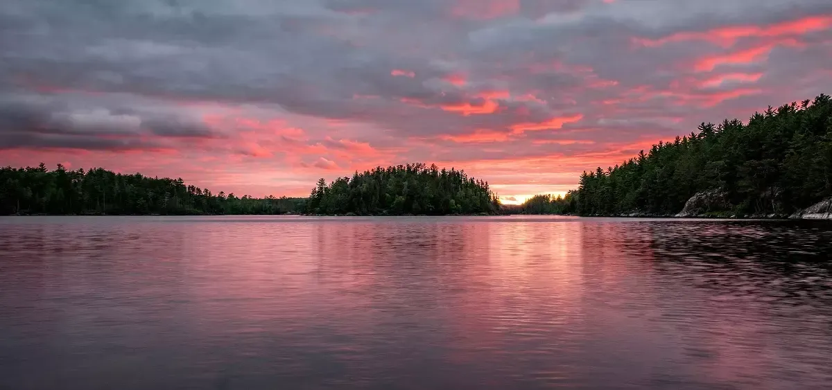 A calm lake reflects a vibrant pink and orange sunset over silhouetted islands and a lush forest shoreline.