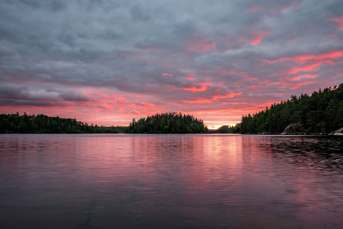 A vibrant, pink and orange sunset glows above a calm lake surrounded by dark, pine-covered islands and shoreline.