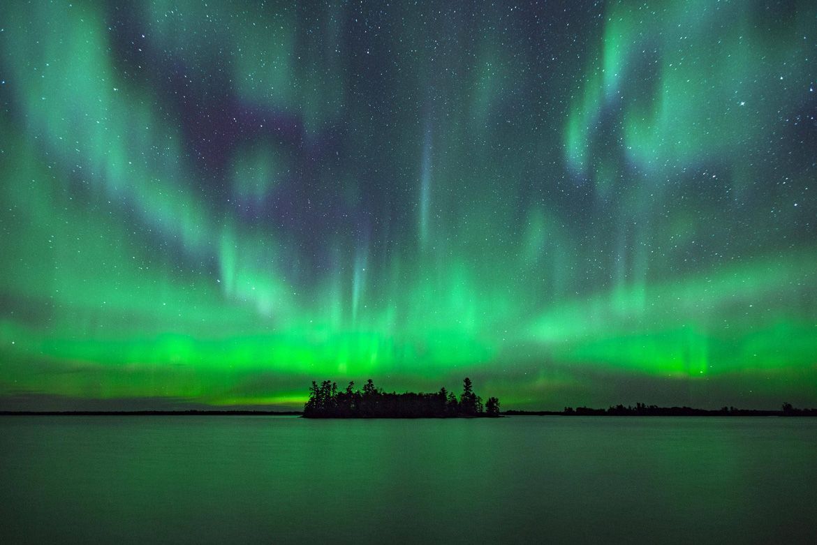 Vibrant green northern lights glow intensely over a dark, silhouetted island surrounded by a calm, reflective lake.