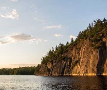 A rocky cliffside covered in pine trees sits at the edge of a calm lake under a soft, golden sunset sky.