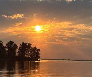 A bright sun sets over a calm lake, casting a golden glow on the water beside a silhouette of trees on a small island.
