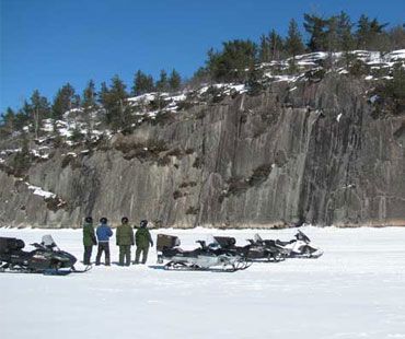 Four people standing beside parked snowmobiles on a frozen, snow-covered lake in front of a rocky cliff.