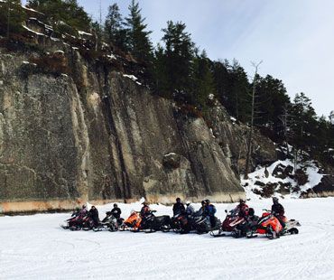 A group of people on colorful snowmobiles parked in a row on a snowy, frozen lake beneath a large, rocky cliff face.