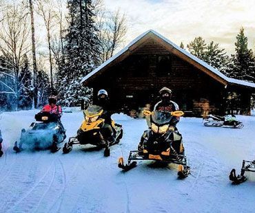 Three people on snowmobiles parked in front of a log cabin in a snow-covered forest.