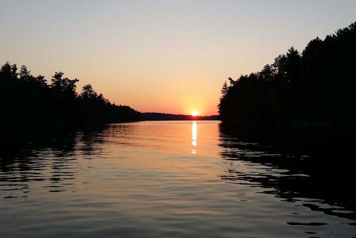 Sunset over a calm lake, framed by dark silhouettes of trees on both sides, with golden light reflecting on the water.