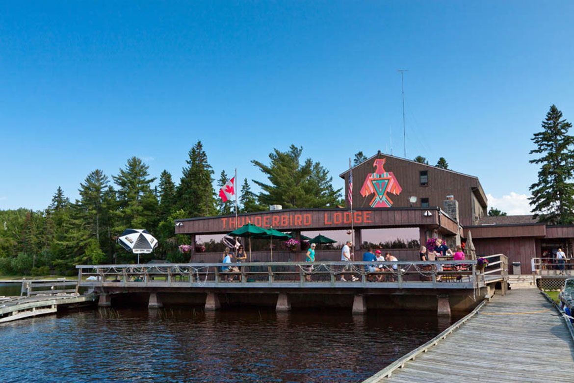 A wooden lodge with a deck over a lake, featuring outdoor seating, a red sign, and surrounding pine trees under blue sky.